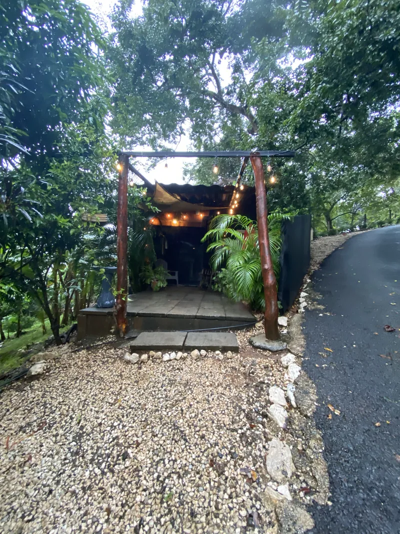 L’entrée abritée du Container Home, encadrée de poteaux en bois et de guirlandes lumineuses, nichée dans les arbres le long du chemin du domaine.
