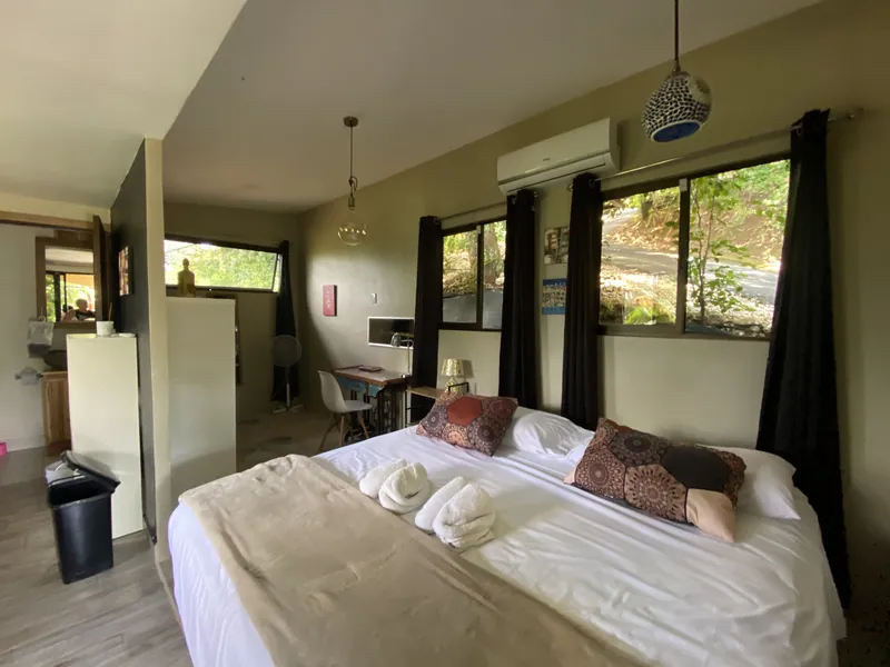 La chambre-studio avec un lit king, des serviettes pliées, un bureau et des fenêtres sur le jardin habillées de rideaux noirs.