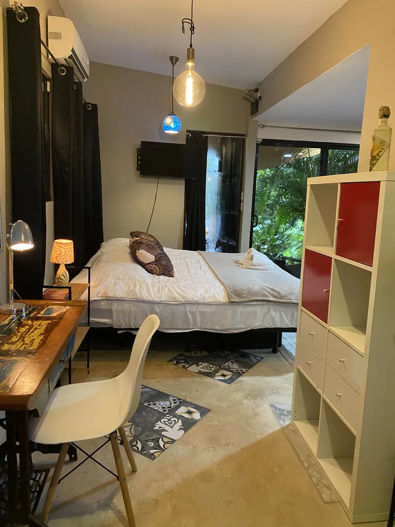 La chambre-studio au crépuscule avec un lit fait, un bureau et une lampe, un téléviseur mural et des portes vitrées coulissantes ouvrant sur le jardin.