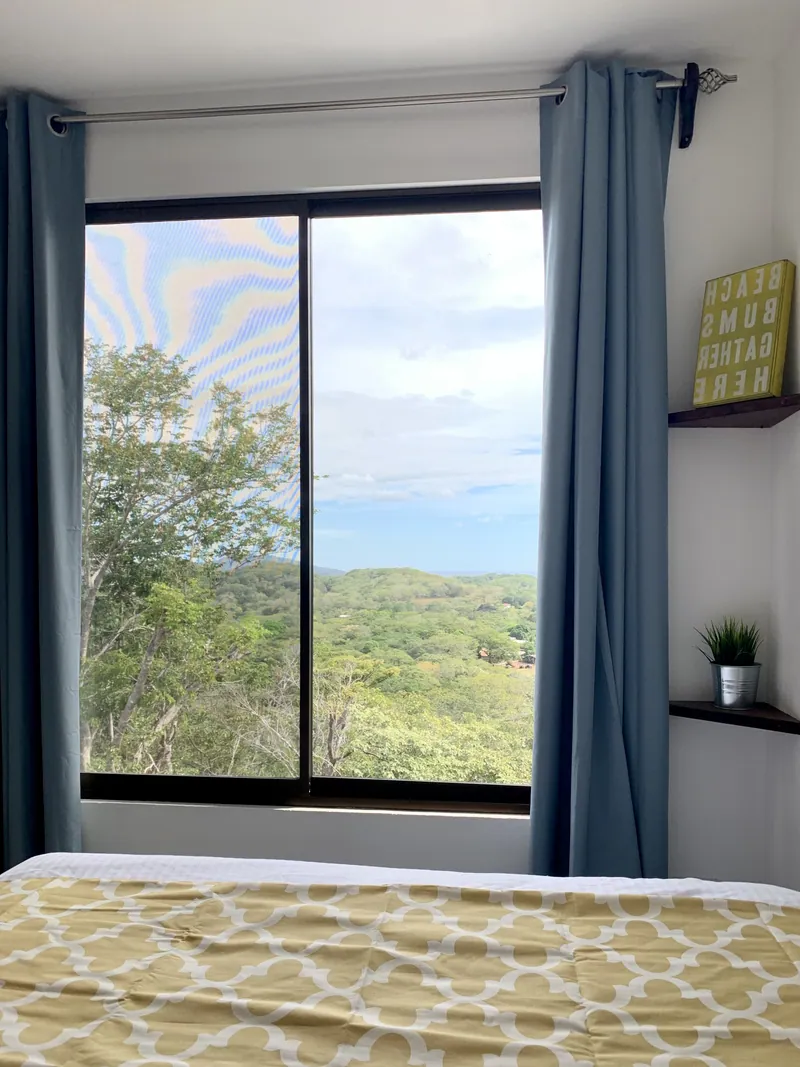 A bedroom window at Villa Alegria framing the valley and distant ocean, with blue curtains